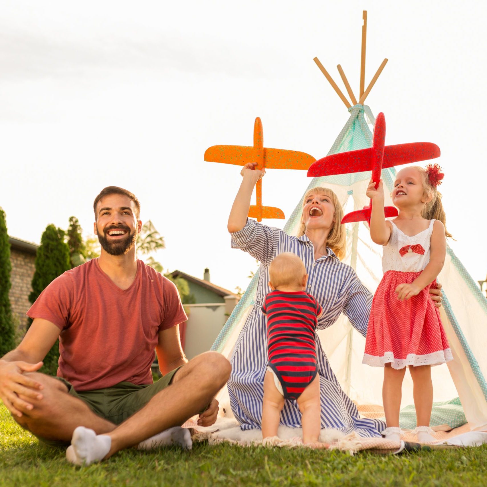 Beautiful young happy family having fun spending sunny summer day camping in the backyard, parents playing with their children, throwing toy airplanes